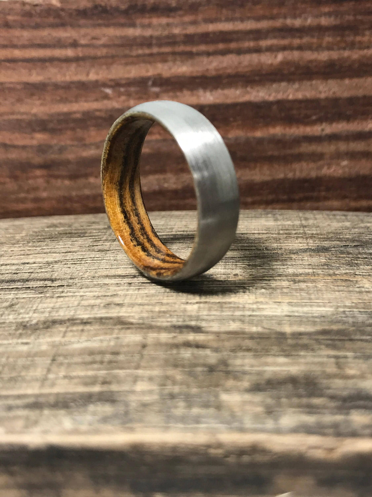 A brushed titanium ring with a dark wood interior stands upright on a rustic wooden board.