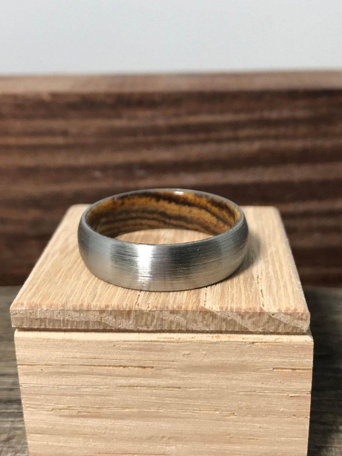 A brushed metal ring with a striped wood interior rests flat on top of a small square wooden box.