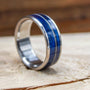 A titanium ring featuring a central blue wood inlay bisected by a thin metal guitar string resting on a wooden surface.