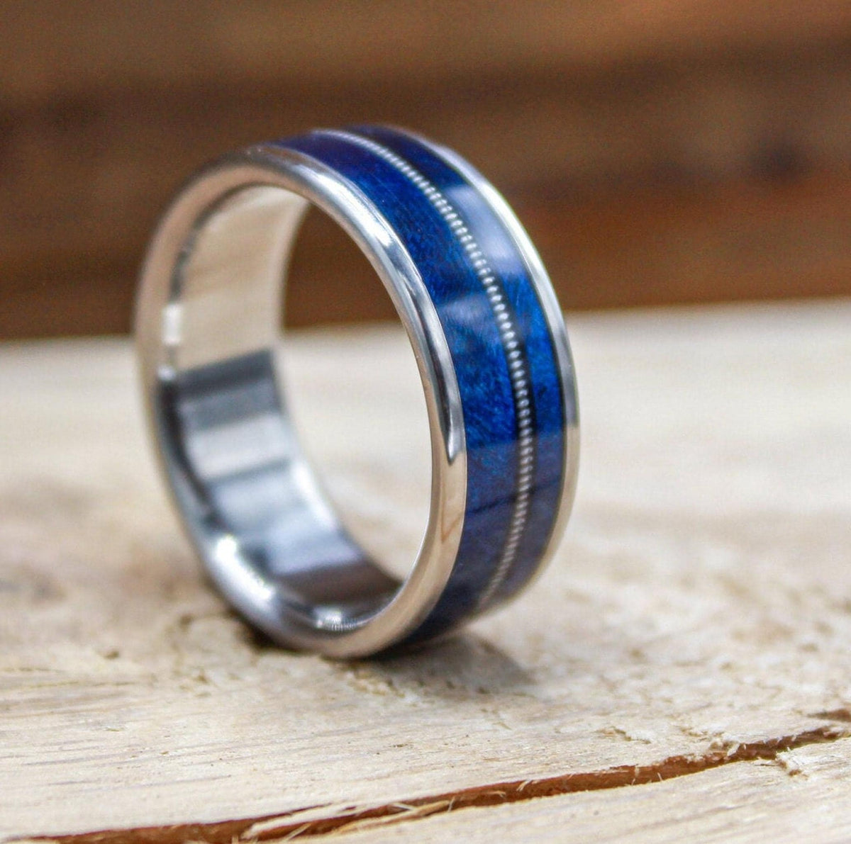 A titanium ring featuring a central blue wood inlay bisected by a thin metal guitar string resting on a wooden surface.