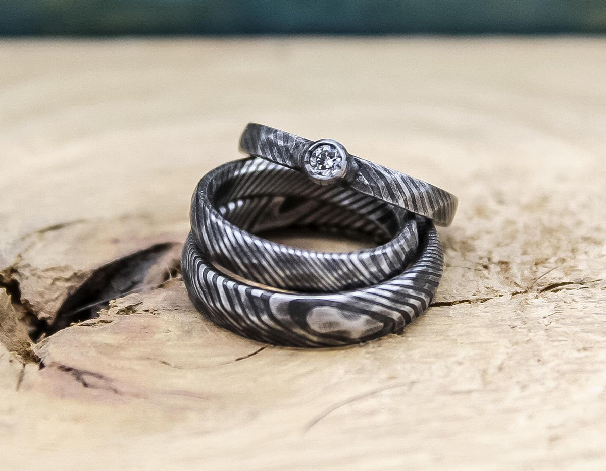 RobandLean's Damascus steel wedding band set with his and hers bands and a diamond engagement ring, featuring intricate craftsmanship.