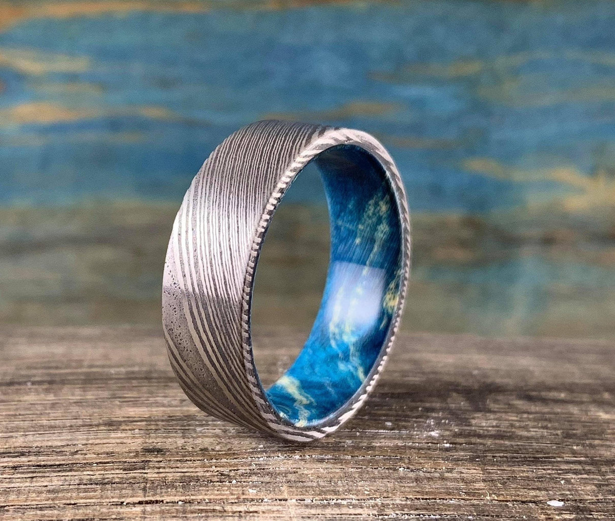 Custom Damascus Steel wedding band with blue Box Elder wood in a natural, close-up view showing the ring's unique wood and steel pattern.