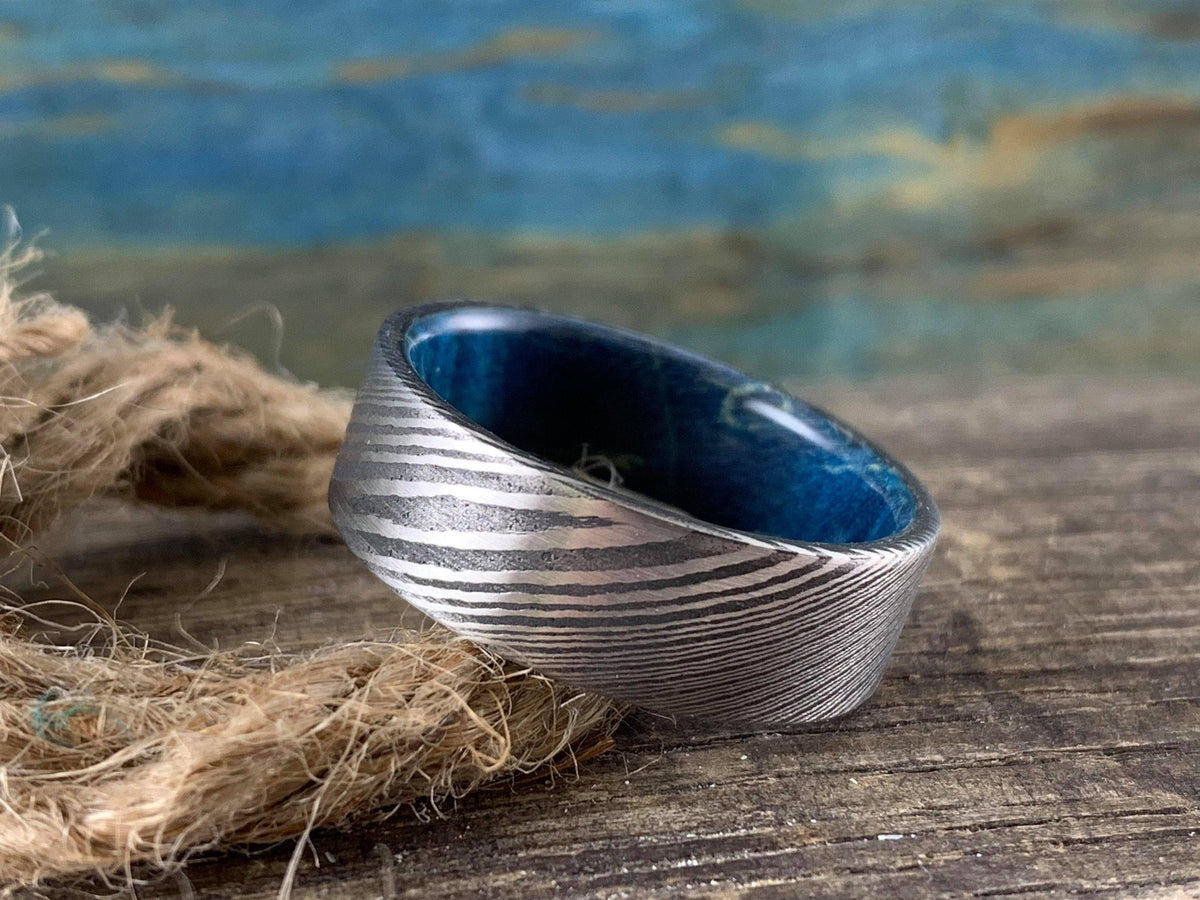 Custom Damascus Steel wedding band with blue box elder wood inlay, crafted by RobandLean's Rings, shown in a close-up view.