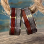 A pair of titanium wedding bands with inlays of redwood and rose quartz, displayed on a wooden surface with a blurred blue background.