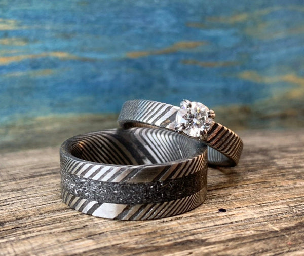 A Damascus steel wedding band with a Gibeon meteorite inlay and a moissanite engagement ring for her, displayed on a wooden surface with a blue textured background.