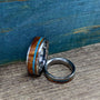 Two titanium rings with inlays of desert ironwood and turquoise, displayed on a wooden surface with a blue textured background.