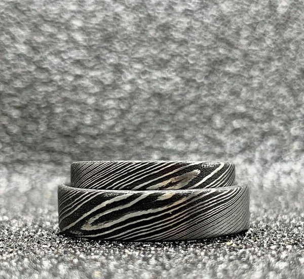 A pair of custom handcrafted Damascus steel wedding bands displayed on a textured surface, showcasing the unique striped pattern of the metal.