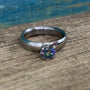 A titanium engagement ring with a prong-set mystic topaz centerpiece, displayed on a wooden surface with a blue item in the background.