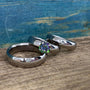 A set of two titanium wedding bands and an engagement ring with a central mystic topaz, displayed on a wooden surface with a blue textured background.