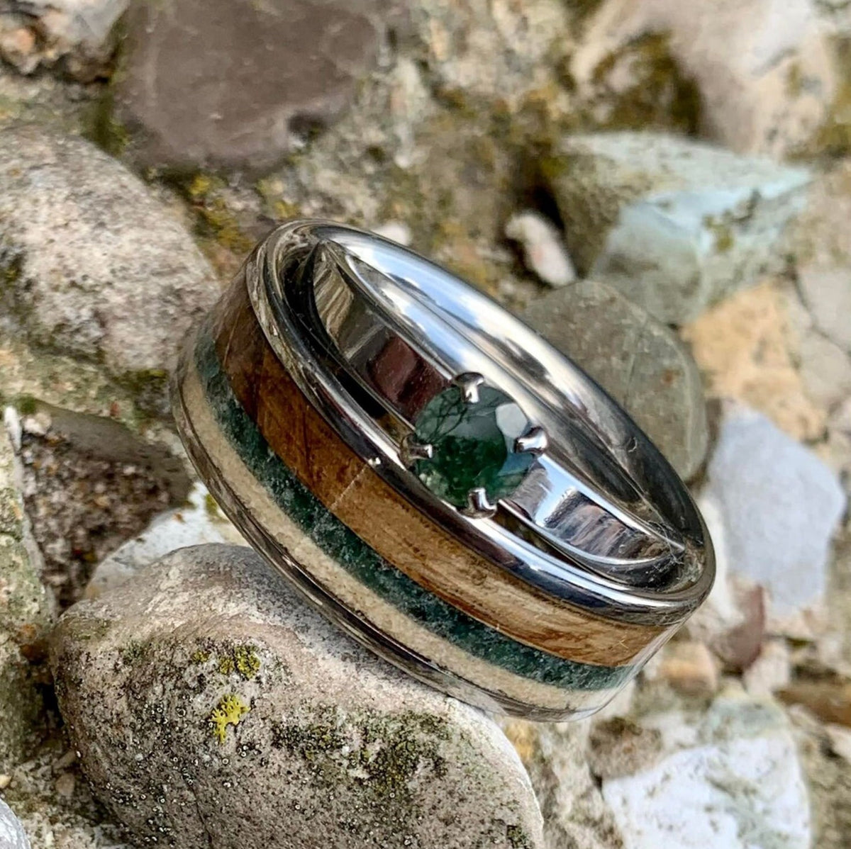 A titanium ring with a segment of English oak wood and a piece of moss agate, alongside a shed deer antler, placed on a rock. alongside a moss agate prong set ring