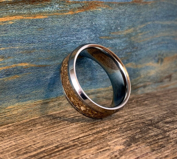 A men's tungsten wedding band with a T-Rex bone inlay, displayed on a textured background.