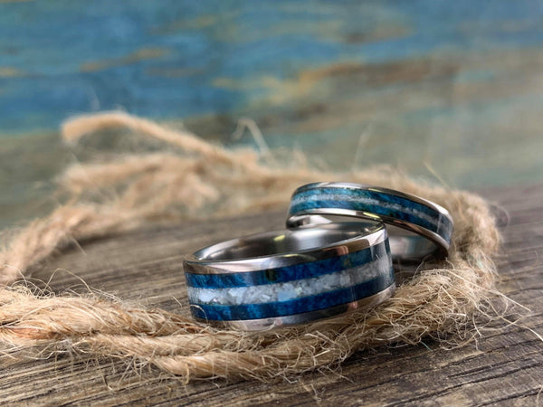 Two titanium wedding bands set with blue wood and pearl inlays, placed on a rope.