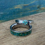 A set of two titanium rings with a blue green moissanite engagement ring placed on top of a band ring, featuring inlay patterns of green goldstone and chrysocolla on a wooden surface with a blurred blue background.