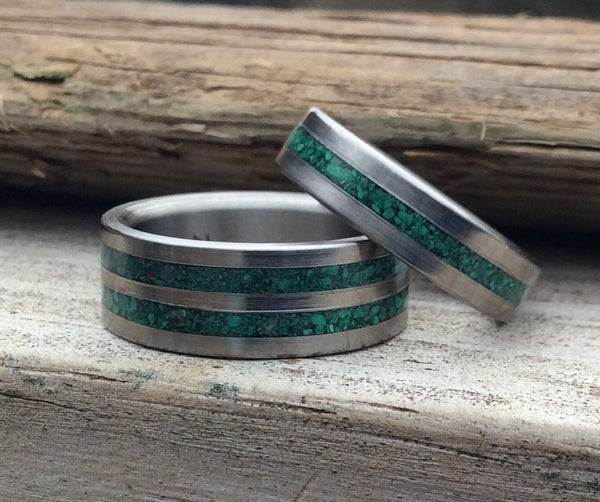 A set of two titanium rings, both with a green malachite stone inlay, placed on a wooden surface.