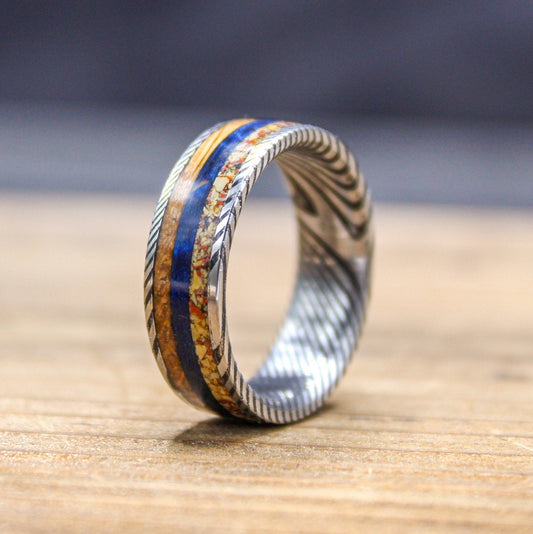 Damascus steel wedding band with dino bone, blue box elder, and whiskey barrel wood in a RobandLean's rings presentation.