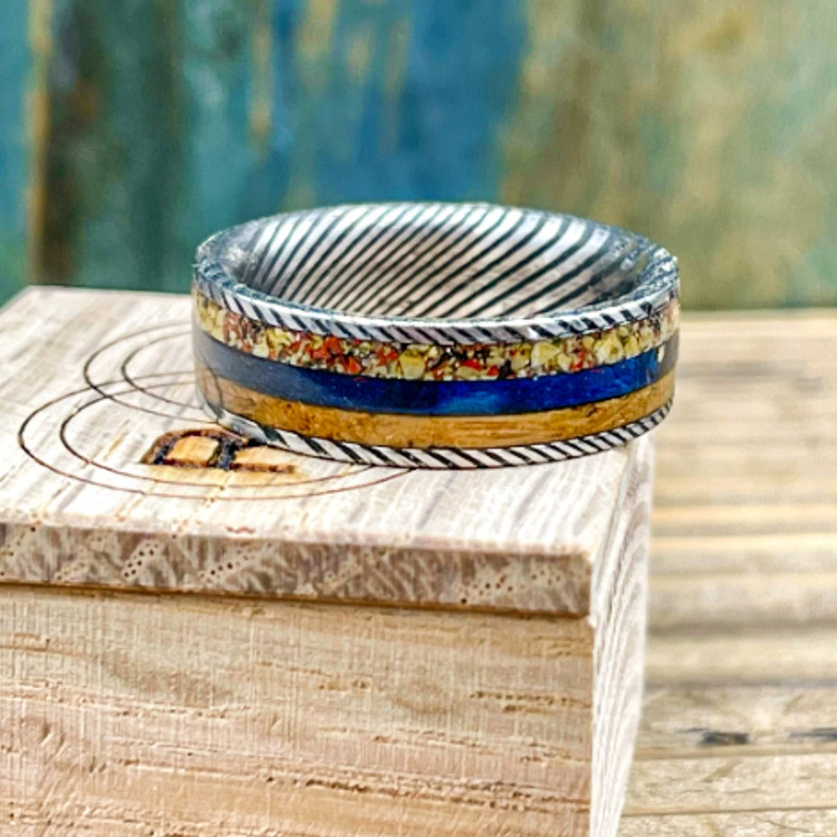 Damascus steel wedding band by RobandLean's with dino bone, blue box elder, and whiskey barrel wood inlay on a white background.