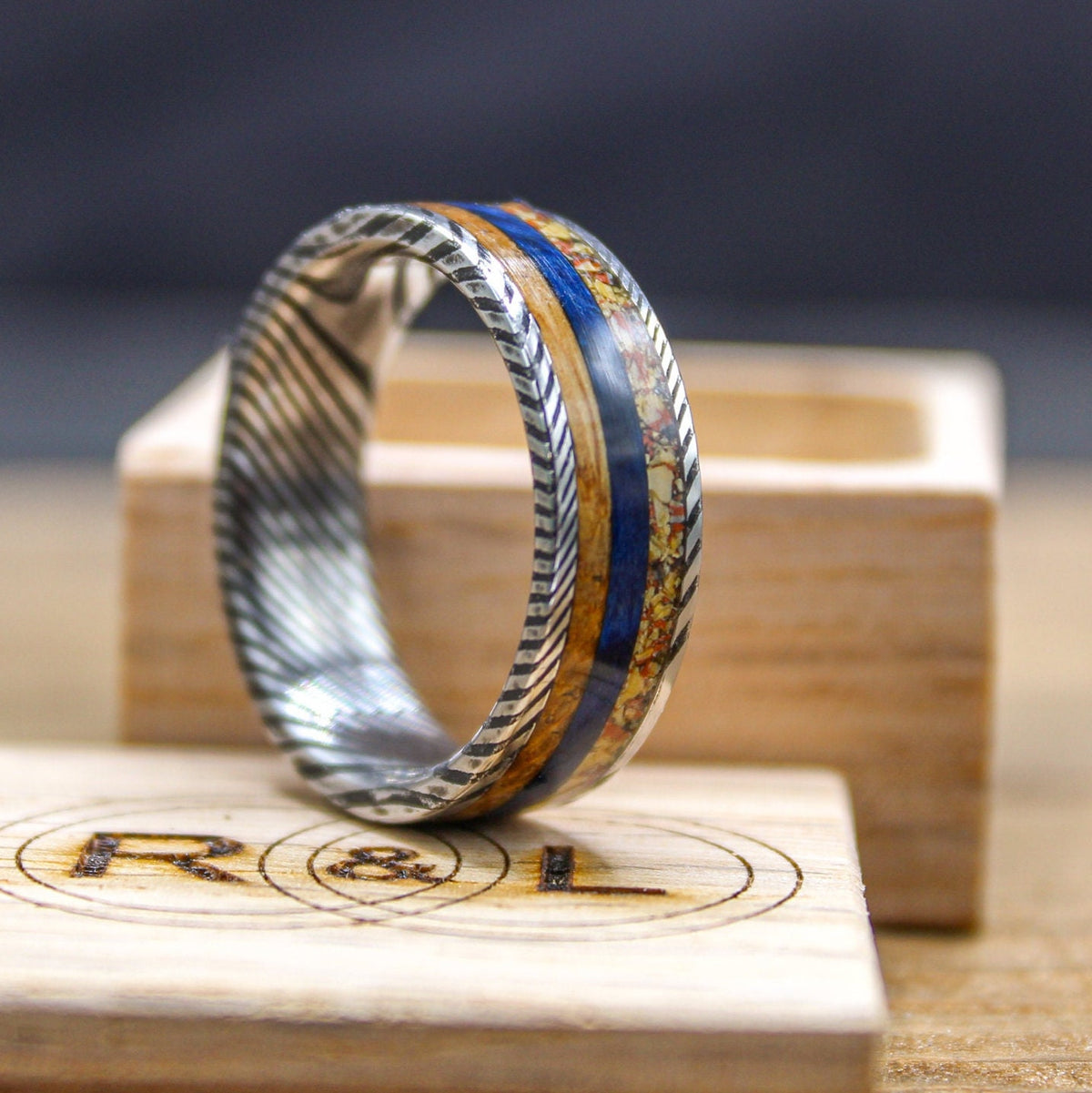 Damascus steel wedding band with dino bone, blue box elder, and whiskey barrel wood accents, handcrafted by RobandLean's rings.